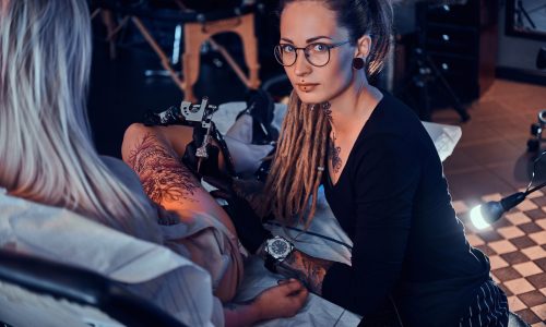 Beautiful creative master with dreadlocks is working on new peace of big leg tattoo for customer.